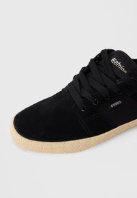 Sapatilhas em camurça negra com sola texturizada bege, atacadores pretos e pequenas etiquetas com o logótipo "etnies" nas laterais e na língua.
