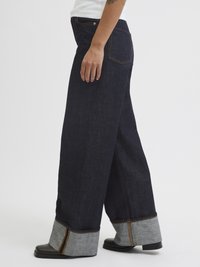 Jeans de perna larga em azul escuro com um punho cinza claro em contraste, apresentando costura laranja e um design clássico de bolso. Textura de denim suave.