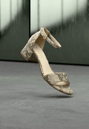 Beige sandal med slangeprint, blokhæl og ankelrem, svævende over en grå overflade med en sløret grøn baggrund.