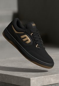 Sapatos de skate pretos com uma textura suave e detalhes em dourado. Apresentam um colarinho almofadado, design com atacadores e uma sola de borracha com acabamento em borracha natural.