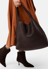 Donna che indossa stivaletti ankle marroni e una gonna ruggine, mentre tiene una grande borsa tote in pelle intrecciata marrone scuro con manici intrecciati.