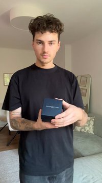 Čierna krabička s textom "TOMMY HILFIGER" držená v rukách. Hladký matný povrch, štvorcový tvar. Pozadie obsahuje svetlú pohovku a zrkadlo.