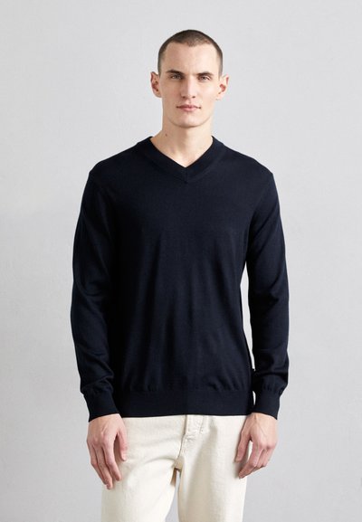 Pull en V marin en tricot doux, avec des manches longues et une taille ajustée, associé à un pantalon de couleur claire.