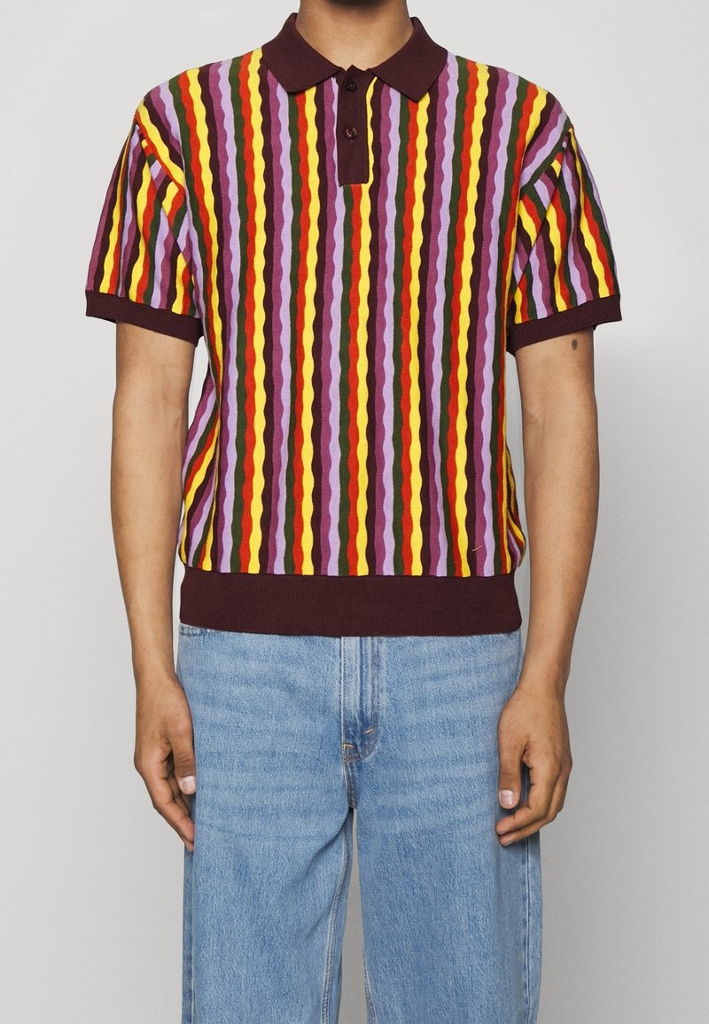 Personne portant un polo à manches courtes avec des rayures verticales ondulées multicolores en violet, jaune, rouge et vert, associé à un jean bleu clair.