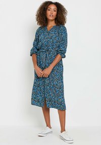 Vestido midi de estampado floral azul con escote en V, cintura ceñida y mangas largas. Combinado con zapatillas blancas, presenta una abertura en la parte delantera.