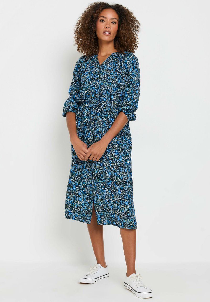 Vestido midi de estampado floral azul con escote en V, cintura ceñida y mangas largas. Combinado con zapatillas blancas, presenta una abertura en la parte delantera.