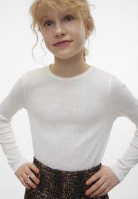 Vit långärmad topp med ett strukturerat mönster, rund halsringning och en fin spetskant. Tillsammans med en brun, mönstrad kjol.