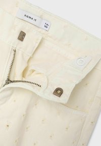 Pantalon en coton couleur crème avec une braguette zippée et un bouton-pression. Intérieur avec un motif texturé et une étiquette de marque visible.