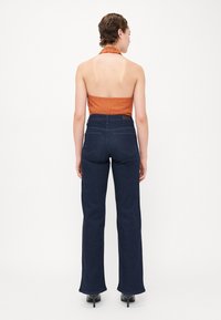 Top z halter-izrezom iz oranžne tkanine z rebrasto teksturo, v kombinaciji z temno modrimi hlačami visokega pasu. Hlače imajo ravne nogavice in zadnji žep.