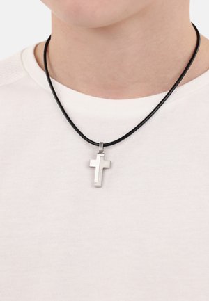 Silberner Metallkreuzanhänger an einem schwarzen Lederband, um den Hals getragen, über einem weißen strukturierten Hemd. Das Kreuz hat ein einfaches, modernes Design.