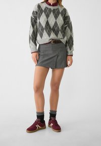 Grauer Argyle-Pullover, dunkelgrauer Minirock mit seitlicher Falte, kastanienfarbene Sneaker und grau-schwarze Socken. Ein brauner Gürtel betont das Outfit.