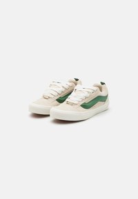 Beige canvas sneakers with green suede accents, round toe, white laces, and textured surfaces, featuring a low-profile design and reinforced heel.