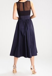 Marineblaues Kleid mit tailliertem Oberteil, durchscheinendem Rückenpanel und ausgestellt knie-langen Rock. Getragen mit metallischen High Heels.