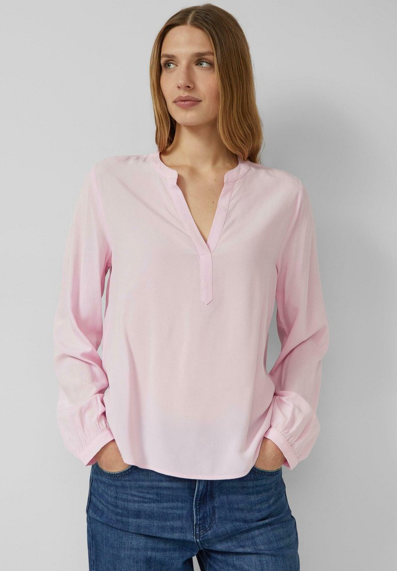 Jasnoróżowa bluzka z długim rękawem o luźnym kroju, z kołnierzem z wycięciem i miękką fakturą. Noszona z niebieskimi dżinsami dla swobodnego stylu.