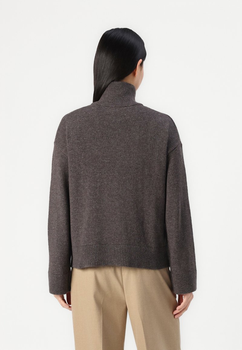 Person mit langen schwarzen Haaren, die einen dunkelgrauen Rollkragenpullover und beige Hose trägt, steht vor einem schlichten weißen Hintergrund.