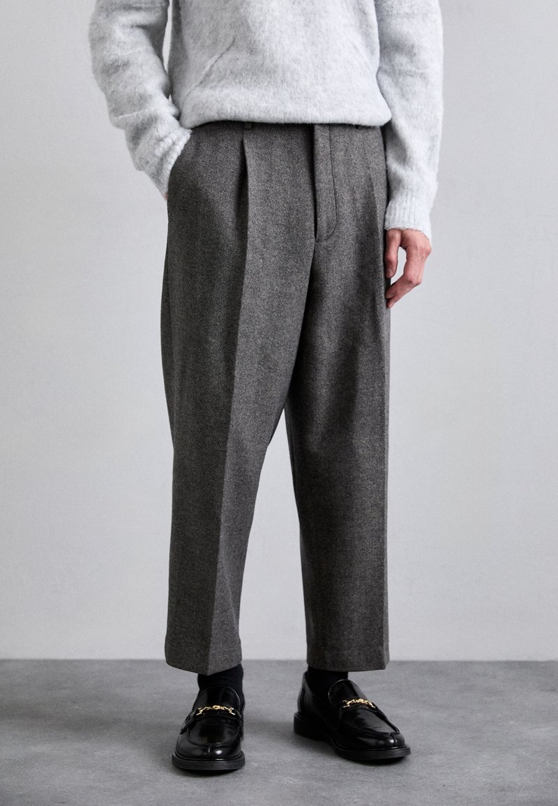 Grå skræddersyede bukser med plisseret front, lavet af et tekstureret stof. Båret med sorte loafers med guldfarvede detaljer og en lys grå sweater.