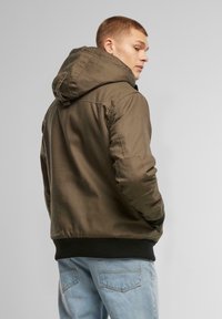Jachetă cu glugă de culoare verde măslinie, cu o textură netedă, manșete și tiv negru ribbat, având cusături vizibile și un design practic.