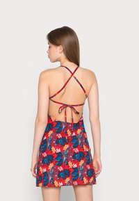 Vestido rojo con flores, que presenta hojas azules y flores naranjas, con un diseño sin espalda y tirantes cruzados atados en la cintura.