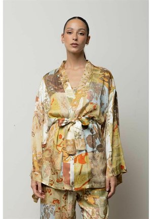 Donna che indossa un abito ampio e decorato, con maniche larghe e cintura annodata in vita, con motivi floreali e astratti in tonalità calde e terrose.