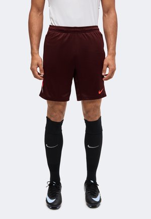 Homme portant un short de sport Nike bordeaux, des chaussettes de football noires avec un logo blanc, des chaussures à crampons noires avec des logos Swoosh bleu clair.