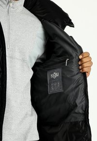 Chaqueta exterior negra con acabado suave; chaleco interior gris con patrón de espiga; bolsillo con cremallera; etiqueta con símbolos de cuidado en el interior.