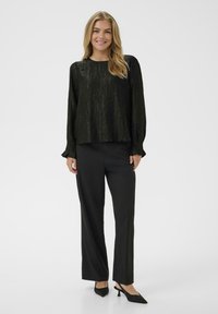 Donna in piedi con una blusa nera a maniche lunghe, texturizzata, pantaloni dritti neri e tacchi a slingback neri a punta, sorridente.