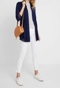 Marinblå cardigan med spetsdetaljer över en randig grå och vit topp, kombinerad med vita slim-fit byxor och vita sneakers; brun axelväska.