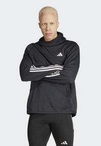 Crna atletska hoodie sa visokim vratom, bijeli Adidas logo i tri bijele pruge na svakoj rukavu, kombinirana s crnim prilagođenim tajicama.