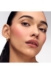 Nærbillede af et ansigt med glat hud, let rouge, neutral læbefarve og mørke, definerede øjenbryn. En guldringeørering fremhæver øret.