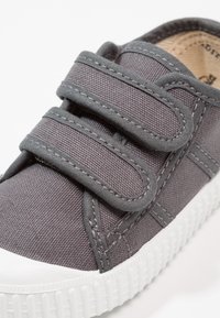 Scarpa in tela grigia con due cinturini regolabili in velcro, suola in gomma bianca, tomaia testurizzata e dettagli di cucitura. Materiale liscio e resistente.