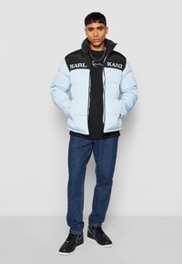 Lys blå puffjakke med svarte skuldre og hvite logoer. Iført over en svart skjorte, kombinert med blå jeans og svarte sneakers.