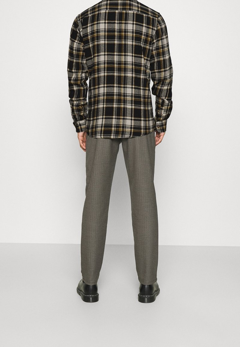 Camisa de franela a cuadros en negro y amarillo combinada con pantalones de espiga en gris. Presenta mangas largas y un diseño de botones, con zapatos negros.