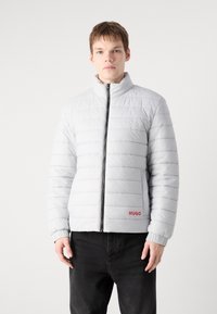 Gai pelēks polsterēts mētelis ar augstu apkakli, priekšējo rāvējslēdzēju, horizontālu dūrienu un sarkano "HUGO" logo kreisajā apakšējā stūrī.