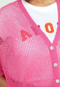 Virkat rosa cardigan med en lös, öppen vävdesign, prydd med pärlknappar och lager på en vit t-shirt med orange tryck.