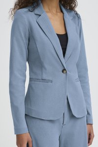 Femeie purtând un sacou albastru deschis, mulat, cu un singur nasture, peste un top negru, asortat cu pantaloni albastru deschis.