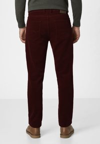 Mann in dunkelroten Cordhosen, braunen Lederschuhen und einem dunkelgrünen Langarmhemd, von hinten vor einem weißen Hintergrund gezeigt.