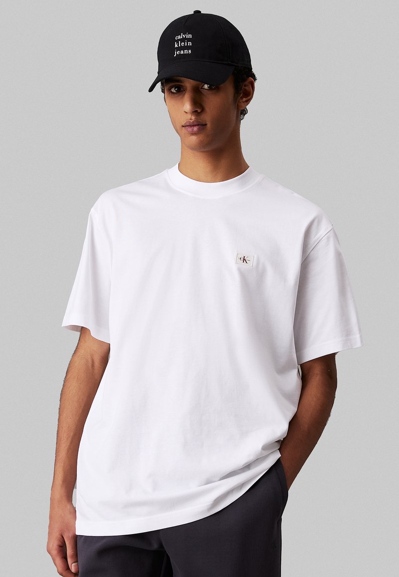 T-shirt bianca in cotone con maniche corte, caratterizzata da una piccola patch con il logo CK sul petto. Abbinata a un cappellino nero con la scritta "calvin klein jeans."