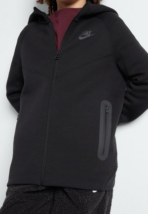 Svart Nike zip-up hoodie med en rymlig huva, texturerad tyg och en diskret logotyp på bröstet. Har en sidoficka med dragkedja för förvaring.
