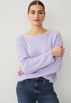 Kvinde med mørkt hår i en lav knold, iført guldringe i ørerne, en lilla ribstrikket sweater og blå jeans, stående med armene over kors.