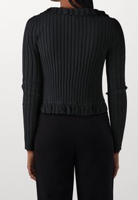 Svart ribbad tröja med långa ärmar, rund halsringning och volangkant. Texturerad tyg med vertikala ränder för extra detalj.
