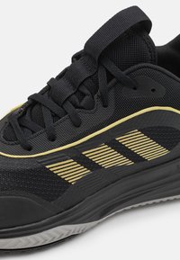 Zapatilla deportiva negra con parte superior de malla transpirable, acentos en amarillos y tres franjas horizontales. Suela de goma texturizada con patrón de tracción.