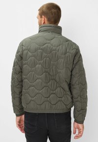 Grøn quiltet pufferjakke med høj krave, prydet med et hexagonalt mønster. Elastiske manchetter og kant, båret med sorte jeans.