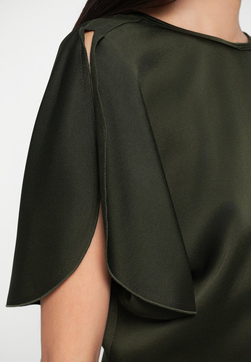 Donkergroene blouse van zachte, vloeiende stof, met uitsnijdingen bij de schouders en delicate stiksels langs de randen.