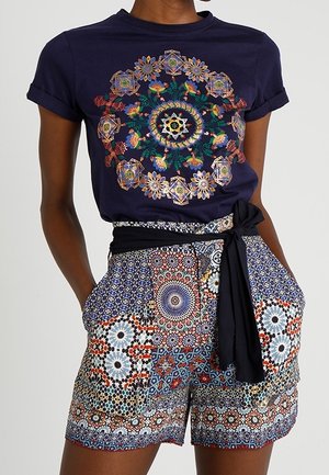 Marineblauw overhemd met kleurrijke bloemige mandala print, gecombineerd met korte broek met geometrische patronen en een zwarte stoffen riem gestrikt in de taille.