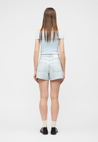 Νέα γυναίκα που στέκεται γυρισμένη με την πλάτη, φορώντας ανοιχτό μπλε τοπ χωρίς ώμους, ανοιχτόχρωμο τζιν σορτς με σποραδικό σχέδιο, λευκές κάλτσες και μαύρα παπούτσια.