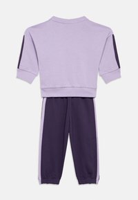 Sweatshirt lila cu mâneci scurte și dungi laterale violete, purtat cu pantaloni joggeri violet cu accente lila și manșete elastice.