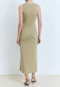 Nainen, joka on pukeutunut hihattomaan, oliivinvihreään midi-mekkoon ja mustiin strap-on-korkokenkiin, seisoo selkä kameraan päin.