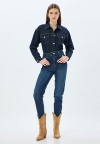 Chaqueta de mezclilla oscura con dos bolsillos en el pecho, cierre de botones y cuello puntiagudo, combinada con jeans oscuros de corte ajustado y botas de vaquero marrones.