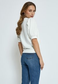 Vit sweatshirt med pösiga ärmar och ribbad nederkant, ihopparad med blå jeans med detaljerade bakfickor. Slät tygstruktur.