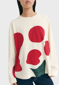 Cream sweatshirt featuring large red abstract shapes and a dark green accent at the bottom. Long sleeves and relaxed fit. Soft fabric texture.